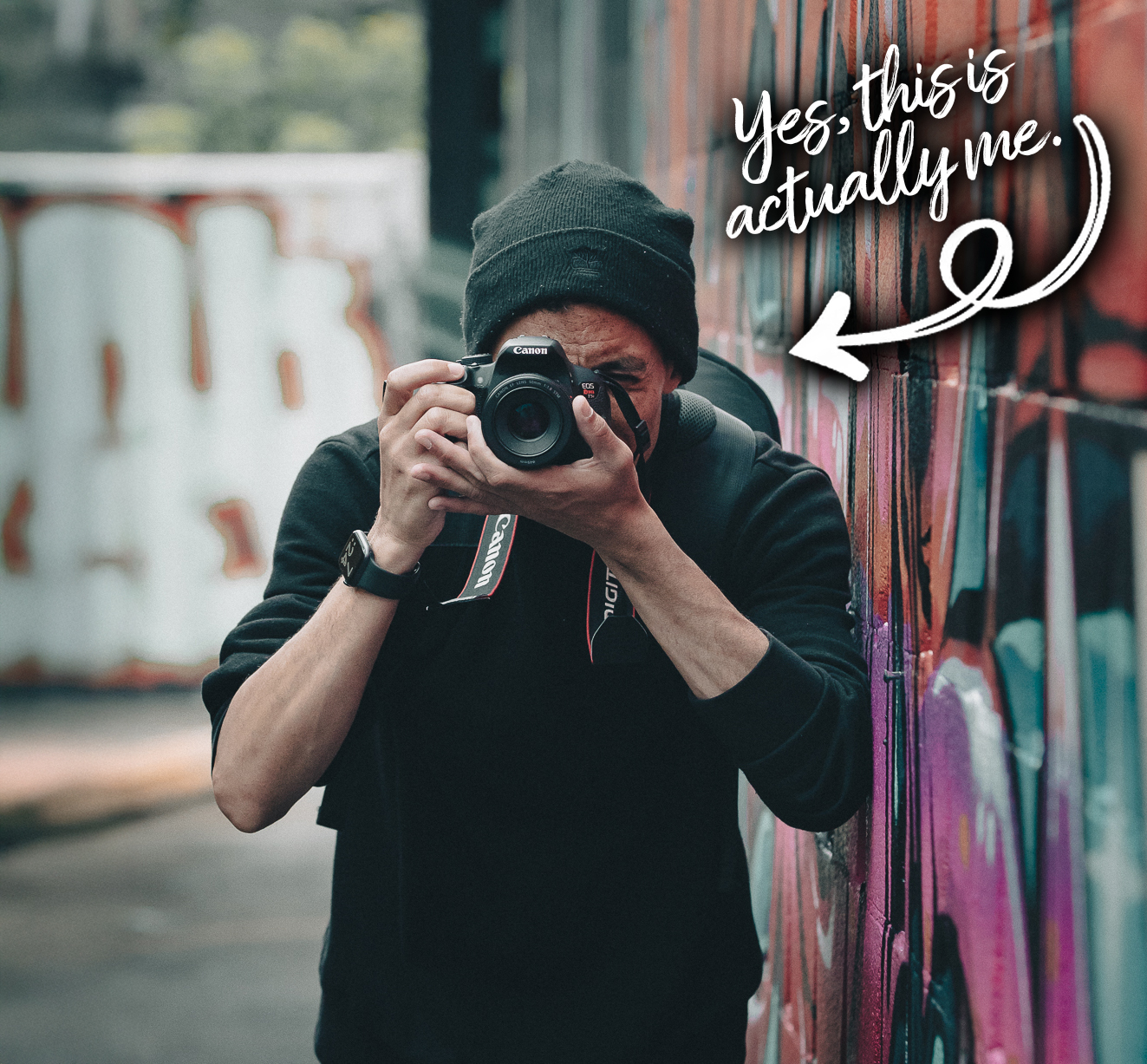 Photographer taking a self-portrait with Canon camera in front of graffiti wall — image includes handwritten text saying “Yes, this is actually me” to introduce the person behind BTTR Marketing’s photography services.